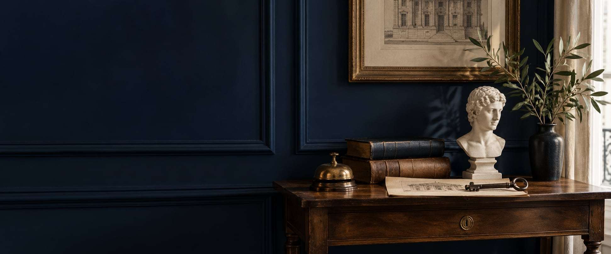 A marble bust, old books, a brass bell and an olive branch on a console table against a navy wall